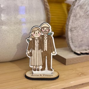Puede incluir: Figura de madera de una pareja, con el texto "Anna & Filippo" en la base. El hombre lleva traje y gorra, la mujer un vestido y chaqueta. La figura est&aacute; sobre una superficie de madera.