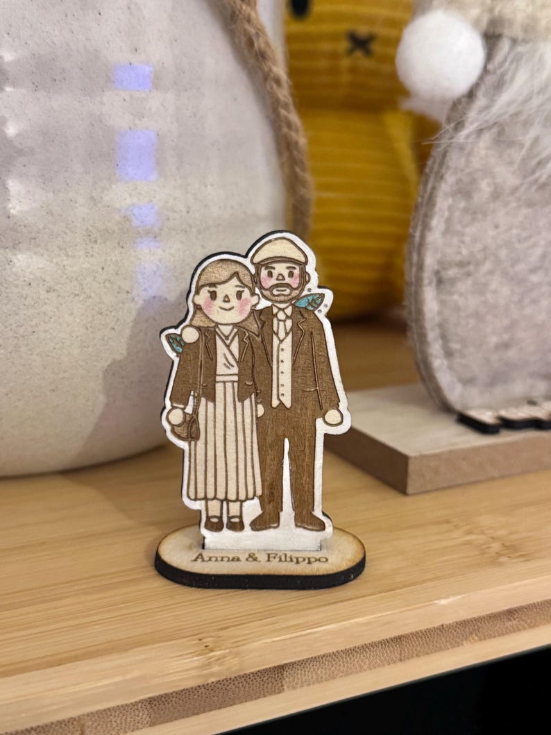 Puede incluir: Figura de madera de una pareja, con el texto "Anna & Filippo" en la base. El hombre lleva traje y gorra, la mujer un vestido y chaqueta. La figura est&aacute; sobre una superficie de madera.