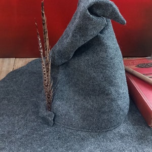 K&ouml;nnte beinhalten: Ein grauer Filz-Zaubererhut mit breiter Krempe und spitzer Krone. Eine braune Feder ist in die Seite des Hutes gesteckt. Der Hut wird auf einer Holzoberfl&auml;che mit rotem Hintergrund pr&auml;sentiert.