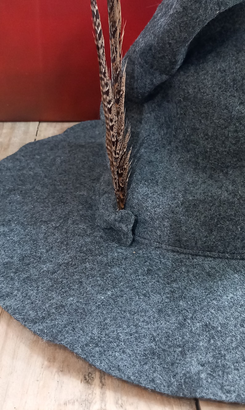 K&ouml;nnte beinhalten: Ein grauer Filzhut mit breiter Krempe, verziert mit einer einzelnen braunen und schwarzen Feder. Der Hut hat ein klassisches Design, das f&uuml;r verschiedene Anl&auml;sse geeignet ist. Die Feder ist in der N&auml;he des Hutes angebracht.