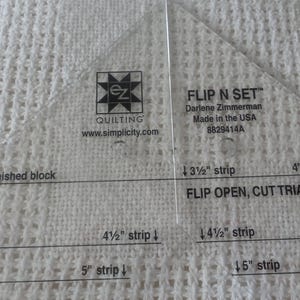 Peut inclure: Une règle de couture en plastique transparent avec des marques pour couper des bandes de tissu et des blocs finis. La règle est intitulée "Flip N Set" et comprend des instructions pour couper des triangles. La règle est conçue pour les projets de courtepointe.
