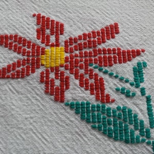 May include: Close-up of a white fabric with a cross-stitched floral design. The flower has red petals, a yellow center, and green leaves and stem. The fabric has a textured, diagonal weave.