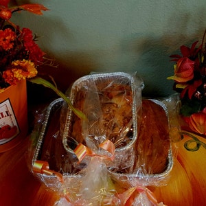 May include: Three loaves of bread wrapped in clear plastic and tied with a ribbon. The loaves are in silver foil pans. The loaves are on a brown surface.