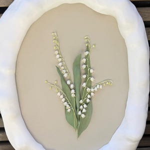 May include: An oval-shaped artwork featuring a botanical arrangement of lily of the valley flowers. The flowers are white with green leaves, set against a beige background. The artwork is framed by a thick, white, cloud-like border.