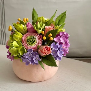 Pode incluir: Um vaso de cerâmica rosa cheio de um buquê de flores artificiais. O buquê inclui flores rosa, verdes e roxas, bem como botões amarelos.