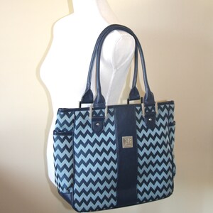 Puede incluir: Un bolso tote azul y blanco con estampado de chevron, ribete de cuero negro y logotipo plateado. El bolso tiene dos asas superiores y cierre de cremallera.