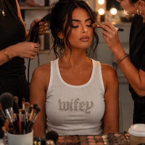 May include: A white ribbed tank top featuring the word wifey written in a gothic-style font across the chest. The text is embellished with small, clear, reflective rhinestones that catch the light, creating a sparkling effect on the fabric.