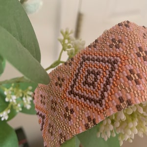 May include: A beaded bracelet with a geometric pattern in shades of pink, brown, and gold.