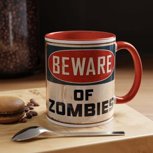 May include: A ceramic coffee mug with a red interior and handle, featuring the words "BEWARE OF ZOMBIES" in a distressed, vintage style. The mug has a white background with a red and blue oval design.