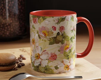 Könnte beinhalten: Eine Keramiktasse mit floralem Muster und rotem Henkel und Innenraum. Die Blumen sind weiß, rosa und gelb auf cremefarbenem Hintergrund. Ein Löffel und ein Macaron liegen auf einer Holzoberfläche.