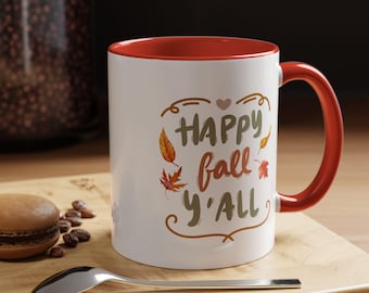 May include: White ceramic mug with a red interior and handle. The mug features the text "HAPPY fall y'all" in brown, orange, and green, with autumn leaf illustrations. The mug is on a wooden surface.