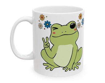 May include: White ceramic mug with a cartoon frog illustration. The green frog is giving a peace sign and winking. The mug is decorated with colorful flowers. The mug has a white handle and is suitable for hot beverages.