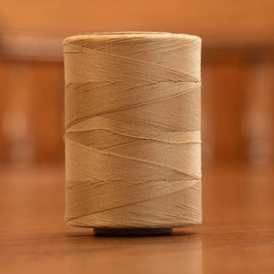 May include: A spool of beige thread wound tightly on a cardboard core.