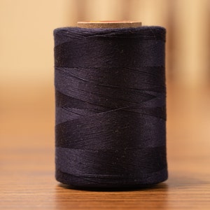 May include: A spool of black thread, tightly wound and cylindrical, stands upright. The thread is a deep, solid black, and the spool has a light brown core. The background is a blurred wooden surface.
