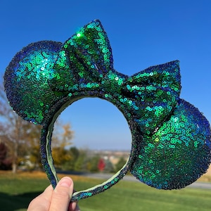 May include: A pair of sequined mouse ears in shades of green and blue. The ears and bow are covered in shimmering sequins, with a green headband. The accessory is held up against a blue sky and green grass background.