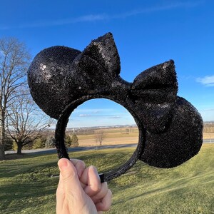 May include: Black sequined mouse ears headband with a matching bow. The ears and bow are covered in small, shiny sequins. The headband is held up against a bright blue sky and green grass.