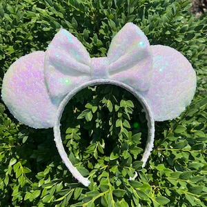 May include: White sequined mouse ear headband with a large bow. The ears and bow are covered in iridescent sequins, creating a shimmering effect. The headband is white and covered in sequins. The accessory is set against a backdrop of green foliage.