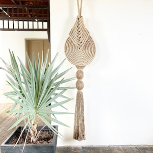 May include: A beige macrame wall hanging with a teardrop shape and a tassel. The hanging is made of natural rope and has a knotted design.