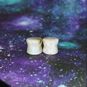 May include: Two white stone ear gauges with brown flecks. The gauges are round and have a smooth surface.