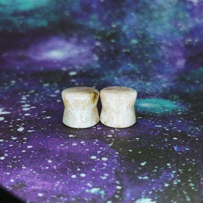 May include: Two white stone ear gauges with brown flecks. The gauges are round and have a smooth surface.