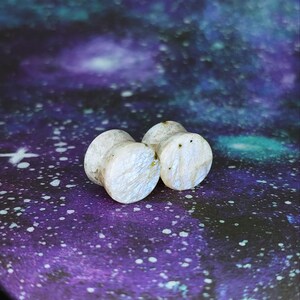 May include: Two white stone ear gauges with gold flecks. The gauges are round and have a smooth, polished surface.