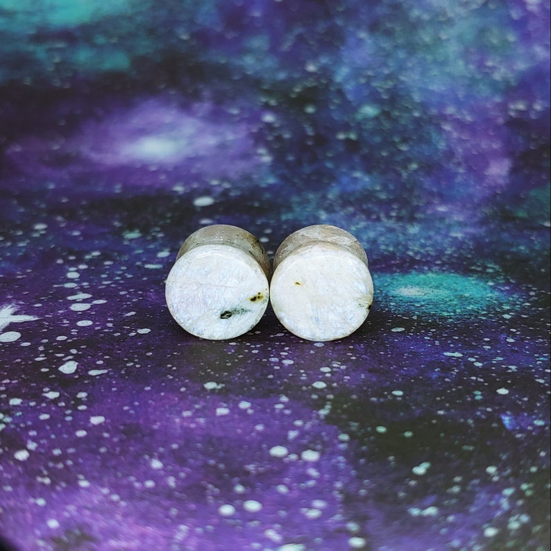May include: Pair of white and grey stone ear gauges. The gauges have a speckled pattern and are round in shape.