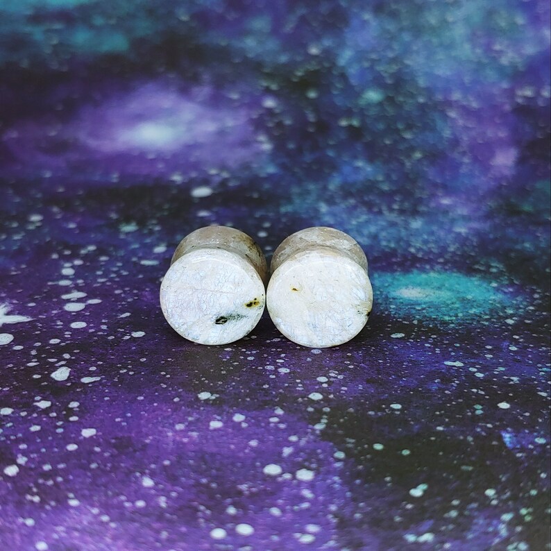 May include: A pair of white wood ear gauges with a subtle shimmer and black flecks.