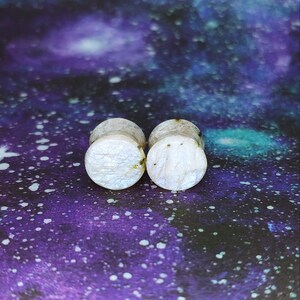 May include: Pair of white and iridescent ear gauges with a speckled pattern.