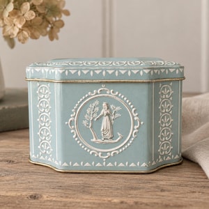 Peut inclure: Une boîte métallique hexagonale bleu clair ornée d'un médaillon central blanc en relief représentant une silhouette debout près d'un arbre. Le récipient présente des bordures et des motifs blancs sur les bords, avec un couvercle assorti et doré.