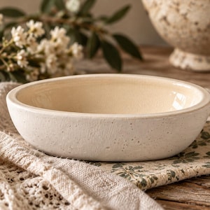 May include: A shallow, oval-shaped ceramic bowl with a textured, off-white exterior and a smooth, glossy cream-coloured interior. The bowl features a speckled finish on the outer surface, showcasing a simple, functional design with a wide rim.