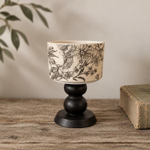 May include: A ceramic bowl with a black and white floral pattern resting on a black wooden pedestal base. The bowl features detailed botanical illustrations of various flowers and leaves, while the stand has a smooth, rounded, stacked design.