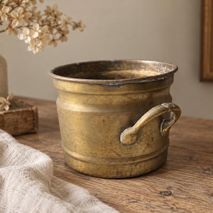 May include: A round, weathered brass pot featuring a wide opening, a slightly tapered body, and two curved side handles. The metal surface displays a dull, aged patina with visible signs of wear, oxidation, and dark metallic discolouration.