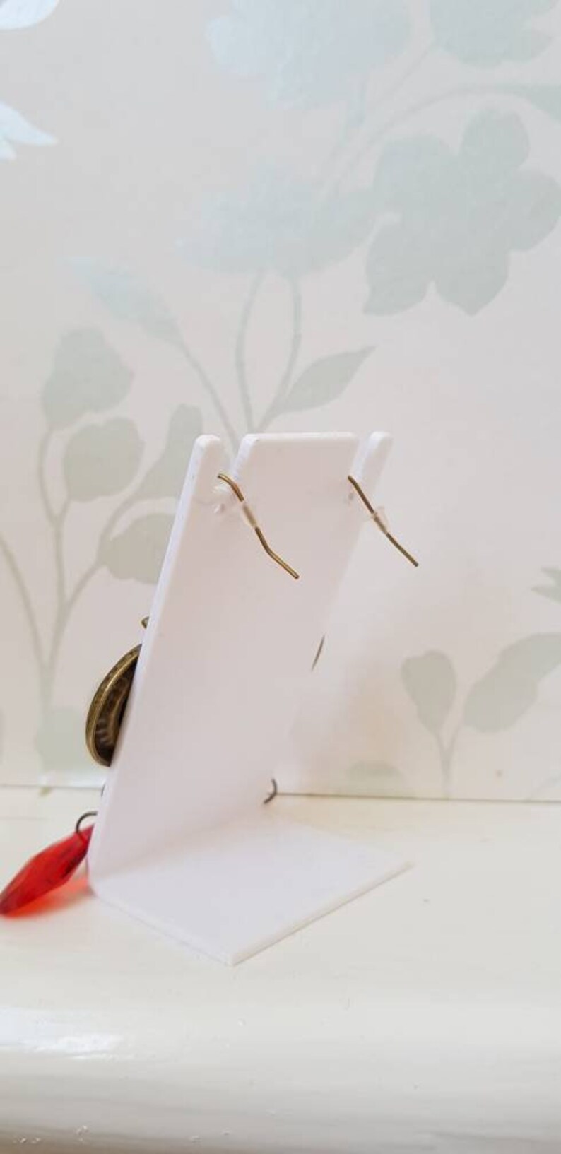 May include: A pair of earrings with a gold-toned metal design and red glass accents. The earrings are displayed on a white stand against a floral background.