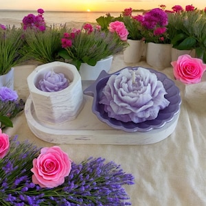 May include: A set of two purple flower-shaped candles in white ceramic holders. The candles are on a white tray with a purple flower-shaped candle in a purple holder.
