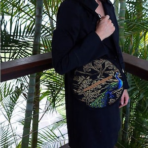 May include: A black crossbody bag with a circular design featuring a peacock and gold accents. The bag is worn by a person standing in front of a palm tree.