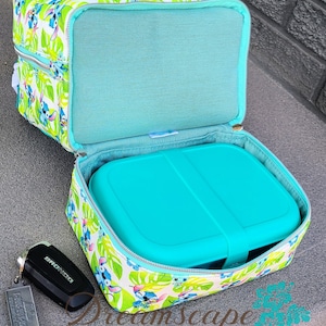 Puede incluir: Una bolsa de almuerzo azul turquesa con fondo blanco y un patr&oacute;n repetido de personajes de Stitch de la pel&iacute;cula de Disney Lilo & Stitch. La bolsa est&aacute; abierta y un contenedor de almuerzo azul turquesa est&aacute; dentro. La bolsa tiene un cierre de cremallera y un bolsillo con cremallera en la parte delantera. La bolsa est&aacute; hecha de tela y tiene un forro suave y acolchado.