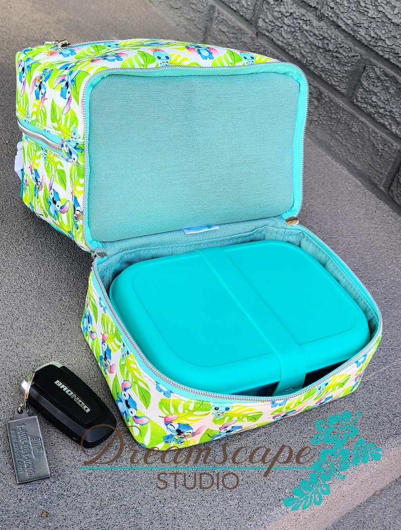 Puede incluir: Una bolsa de almuerzo azul turquesa con fondo blanco y un patr&oacute;n repetido de personajes de Stitch de la pel&iacute;cula de Disney Lilo & Stitch. La bolsa est&aacute; abierta y un contenedor de almuerzo azul turquesa est&aacute; dentro. La bolsa tiene un cierre de cremallera y un bolsillo con cremallera en la parte delantera. La bolsa est&aacute; hecha de tela y tiene un forro suave y acolchado.