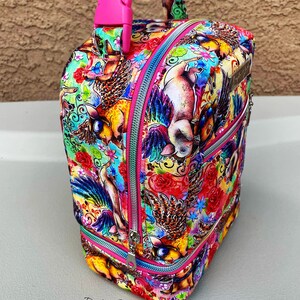 Puede incluir: Una bolsa de almuerzo rosa y plateada con un estampado colorido de cerdos voladores y flores. La bolsa tiene una cremallera rosa y un asa rosa con una hebilla.