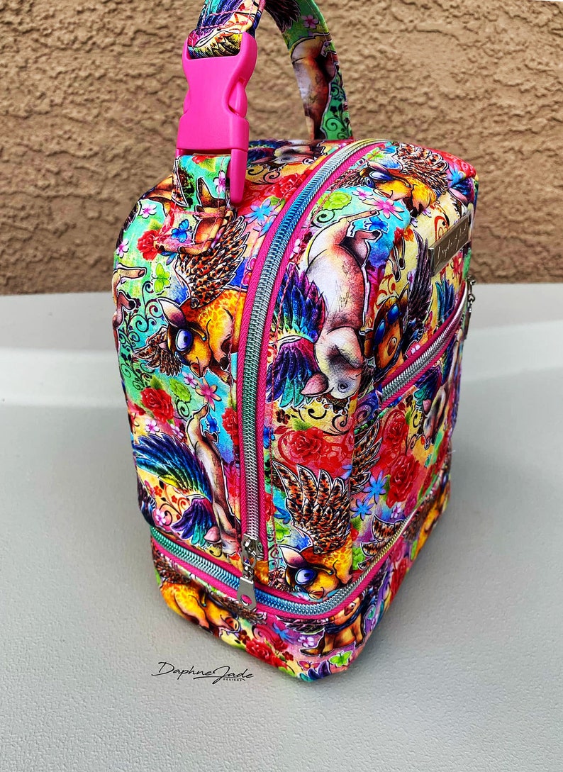 Puede incluir: Una bolsa de almuerzo rosa y plateada con un estampado colorido de cerdos voladores y flores. La bolsa tiene una cremallera rosa y un asa rosa con una hebilla.