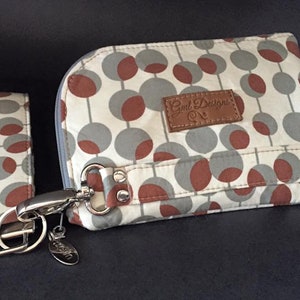 Pode incluir: Uma pequena bolsa de tecido branco e cinza com um remendo de couro marrom que diz "Gnd Designs" e um z&iacute;per prateado. A bolsa tem um clipe prateado e uma al&ccedil;a de pulso de tecido combinando com um chaveiro prateado.