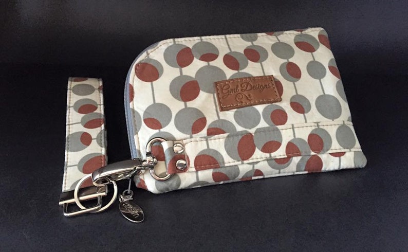 Pode incluir: Uma pequena bolsa de tecido branco e cinza com um remendo de couro marrom que diz "Gnd Designs" e um z&iacute;per prateado. A bolsa tem um clipe prateado e uma al&ccedil;a de pulso de tecido combinando com um chaveiro prateado.