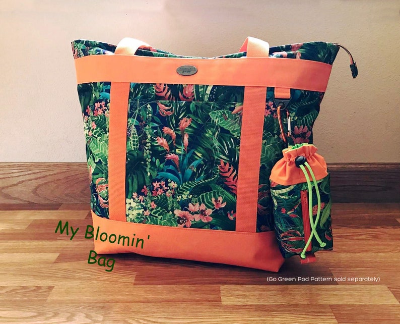 Pu&ograve; includere: Una grande borsa tote con una stampa floreale tropicale, con foglie verdi e fiori rosa, accentuata da finiture e cinghie arancioni. Include una pochette con coulisse abbinata. Il testo "My Bloomin' Bag" &egrave; visibile.