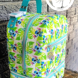Puede incluir: Una mochila turquesa y blanca con un estampado tropical que presenta un personaje de dibujos animados. La mochila tiene dos compartimentos con cremallera y un asa superior. El estampado presenta un personaje azul con orejas grandes y una flor amarilla.