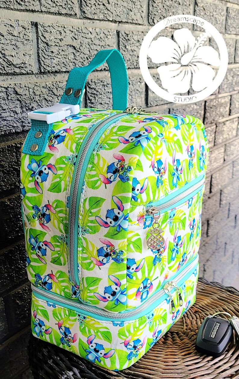 Puede incluir: Una mochila turquesa y blanca con un estampado tropical que presenta un personaje de dibujos animados. La mochila tiene dos compartimentos con cremallera y un asa superior. El estampado presenta un personaje azul con orejas grandes y una flor amarilla.