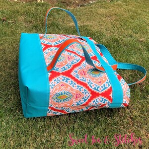 Pu&ograve; includere: Una grande borsa tote con un vivace motivo paisley in rosso, arancione e blu. La borsa presenta lati e manici blu turchese, con accenti arancioni. La borsa &egrave; appoggiata su un prato verde.