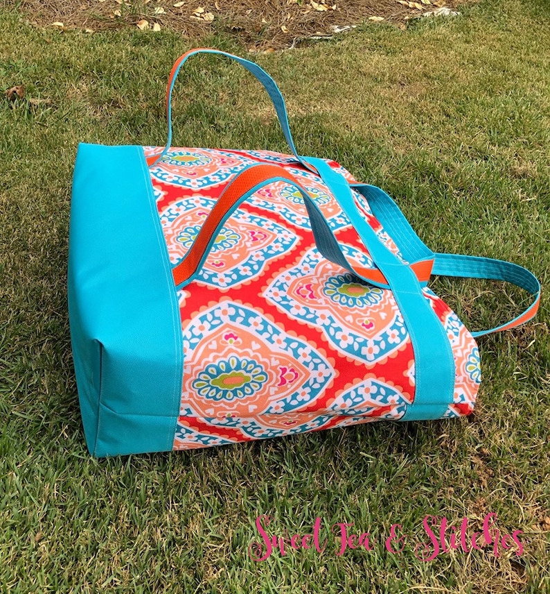 Pu&ograve; includere: Una grande borsa tote con un vivace motivo paisley in rosso, arancione e blu. La borsa presenta lati e manici blu turchese, con accenti arancioni. La borsa &egrave; appoggiata su un prato verde.