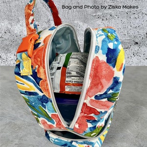 Puede incluir: Una bolsa de almuerzo blanca con un estampado floral colorido. La bolsa est&aacute; abierta y tiene un cierre de cremallera. La bolsa est&aacute; llena de un recipiente azul y un paquete de comida.