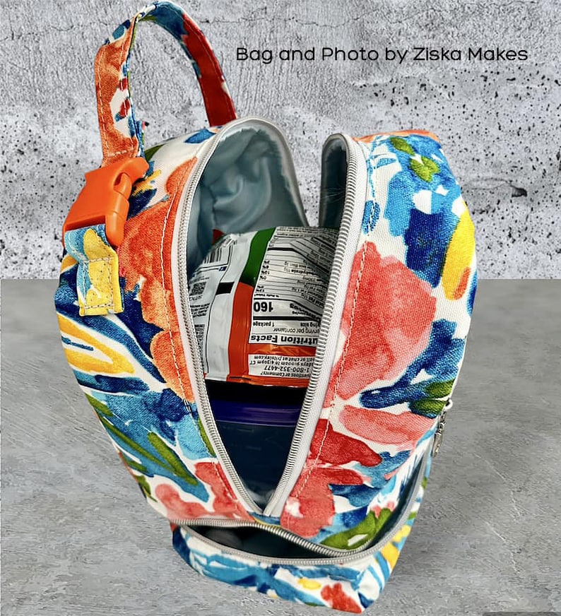 Puede incluir: Una bolsa de almuerzo blanca con un estampado floral colorido. La bolsa est&aacute; abierta y tiene un cierre de cremallera. La bolsa est&aacute; llena de un recipiente azul y un paquete de comida.