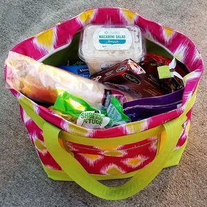 Pu&ograve; includere: Una borsa tote colorata con un motivo ikat rosa, giallo e bianco. La borsa ha finiture e manici verde lime. All'interno della borsa ci sono vari alimenti, tra cui un contenitore di insalata di maccheroni e un pane.