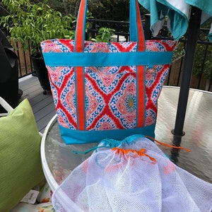 Pu&ograve; includere: Una borsa tote colorata con manici e finiture arancioni e un tessuto fantasia rosso, blu e pesca. Include due sacchetti a coulisse in rete bianca. Il testo "Sweet Tea & Stitches" &egrave; visibile.